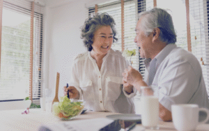 Two elderly people sit at a table with a bowl of fresh salad, glasses of milk, and cups, enjoying a meal in a bright room with wooden blinds.
